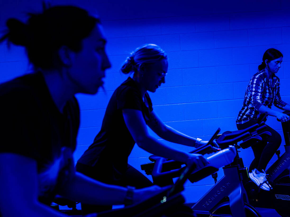 Three students exercise on stationary bikes in a dimly lit room bathed in blue light.